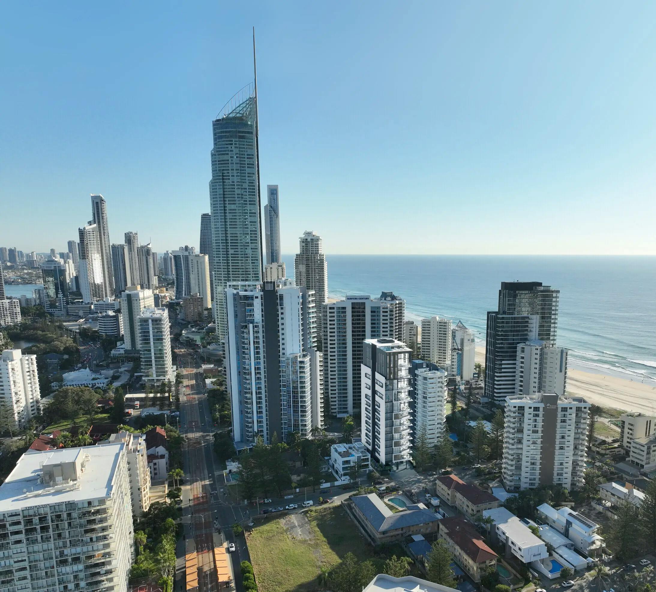 A panoramic view of a coastal city with tall skyscrapers and residential buildings. The cityscape extends along a sandy beach with the ocean in the background. The sky is clear, indicating a sunny day. Notable is a prominent, slender skyscraper on the left.