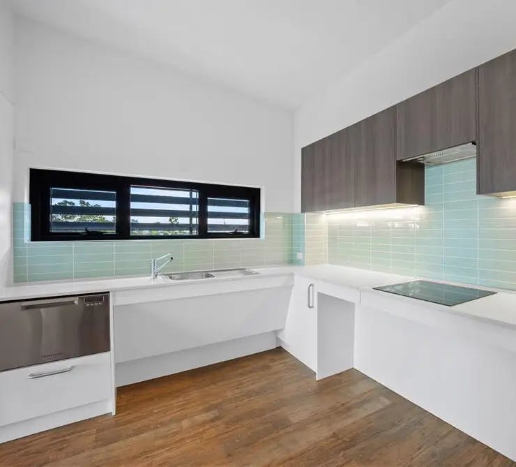 A brightly lit modern kitchen with white countertops, dark wooden upper cabinets, and light green tile backsplash. It features a stainless-steel sink under a black-framed window and an electric stovetop with built-in stove vent under-cabinet lighting. The flooring is wooden.