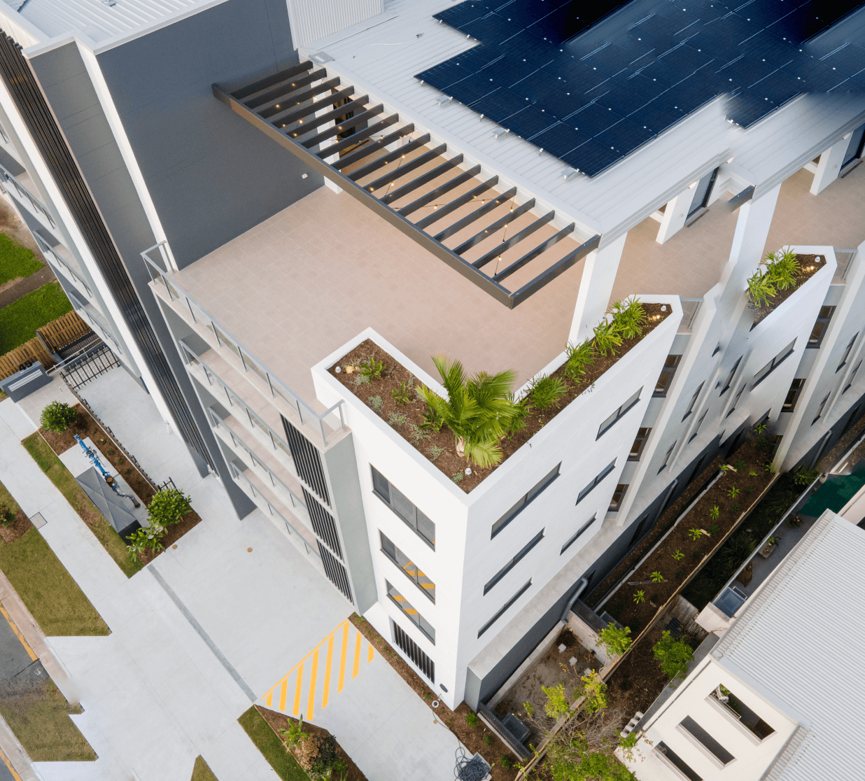 Angled aerial perspective photo of an off white coloured modern residential 6 story building during the day. There are garden beds around the foot of the building and on a roof terrace seen at the centre of the photo. There are balconies at the front of the building, with steel framed glass railings. There are solar panels on the terrace rooftop and the terrace and balconies have cream coloured tiles. In the bottom left of the photo there is a driveway which leads to a carpark out of sight. There is also a concrete walkway across the entrance to the carpark, with a warning strip painted on it. Some plant equipment can be seen in the garden bed adjacent to the driveway. There are wooden paled fences up two sides of the perimeter of the block. There are steel bar fences dividing footpaths from garden beds.